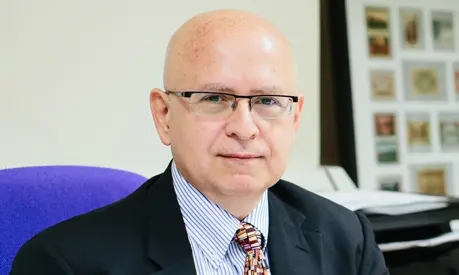 A man in a suit sitting on a purple chair with framed pictures on the wall behind.