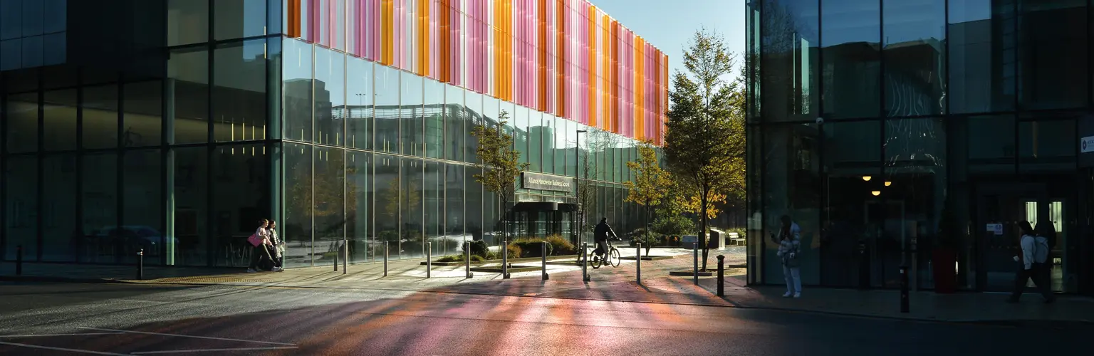 Exterior view of Alliance Manchester Business School with modern glass architecture and colorful vertical panels reflecting light onto the pavement.