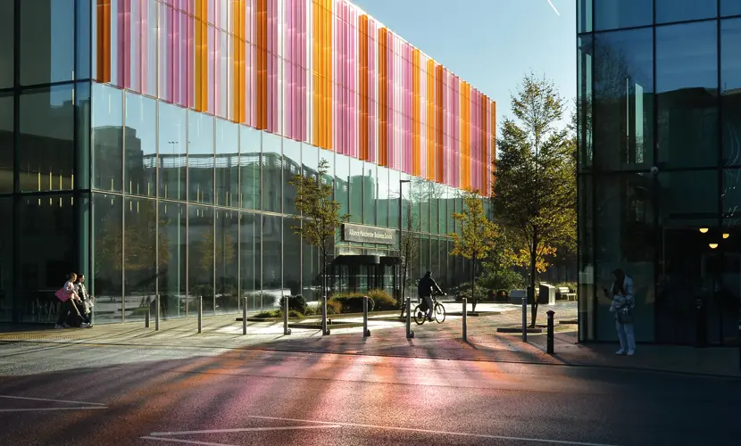Exterior view of Alliance Manchester Business School with modern glass architecture and colorful vertical panels reflecting light onto the pavement.