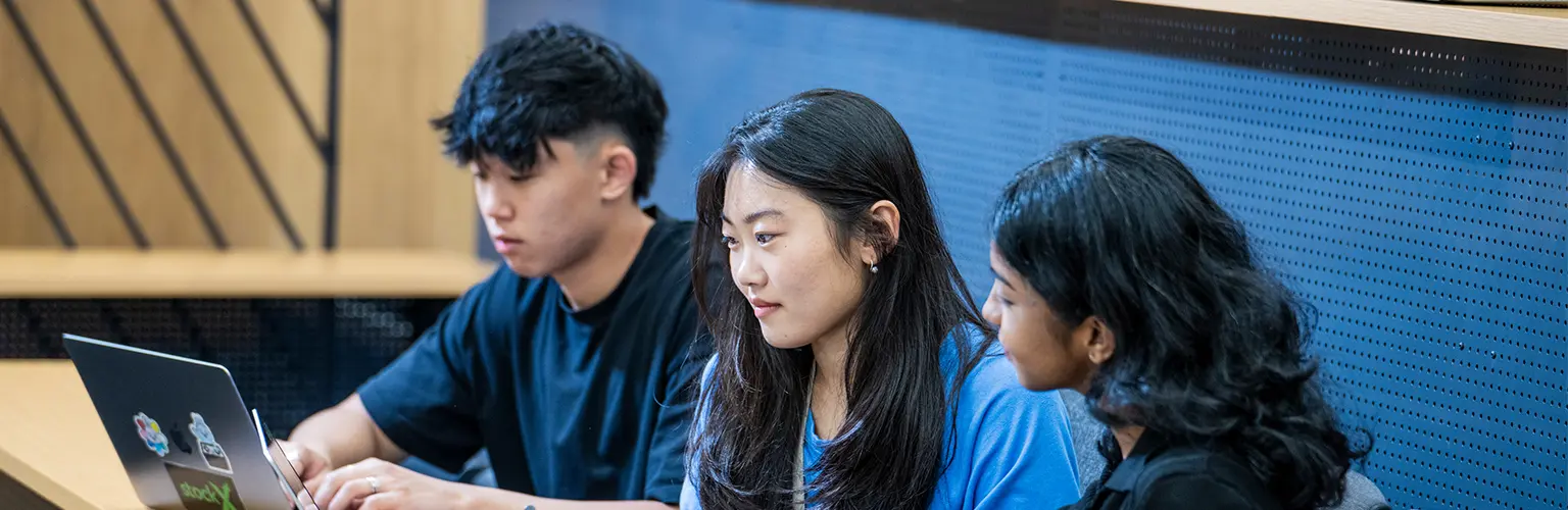 Three students sitting in a lecture hall, working together with laptops and notebooks during class.