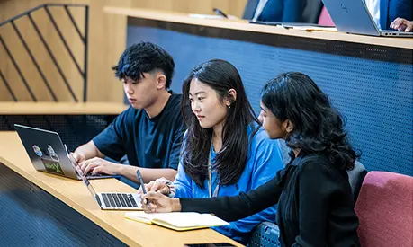 Three students sitting in a lecture hall, working together with laptops and notebooks during class.
