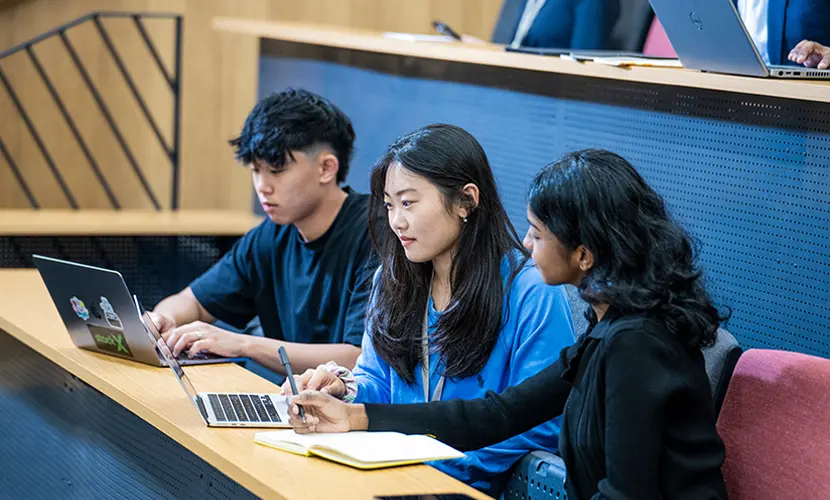 Three students sitting in a lecture hall, working together with laptops and notebooks during class.