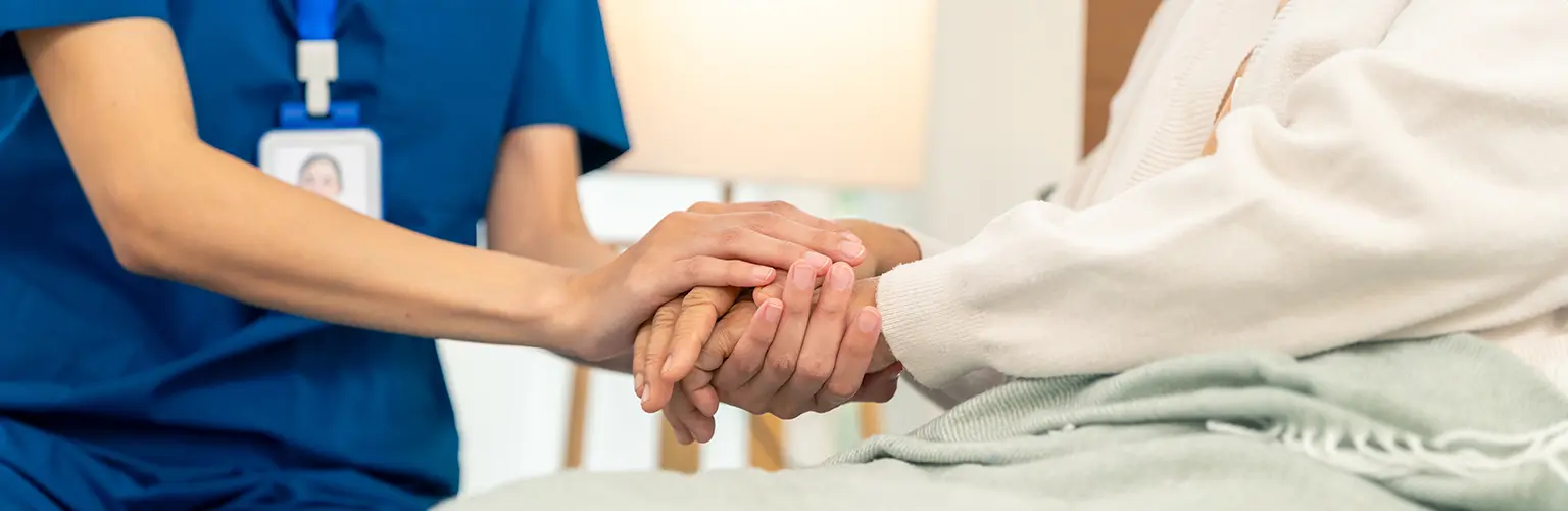 Woman caregiver holding hands with senior woman who is sitting in bed.