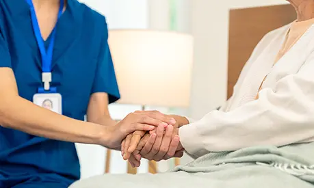 Woman caregiver holding hands with senior woman who is sitting in bed.