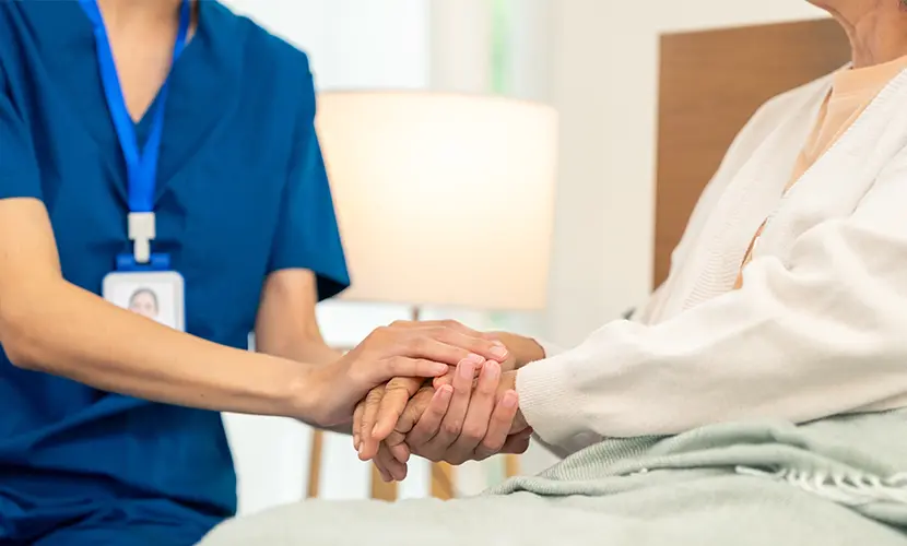 Woman caregiver holding hands with senior woman who is sitting in bed.