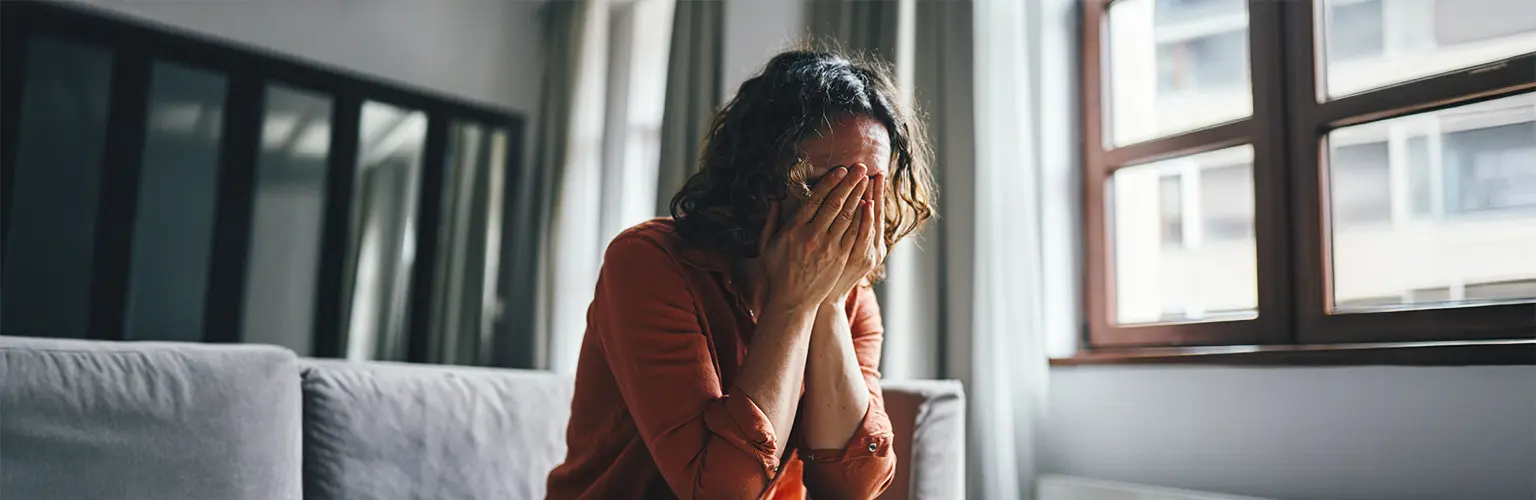 A model poses as a woman covering her face while sitting in a living room, conveying feelings of sadness or emotional stress.