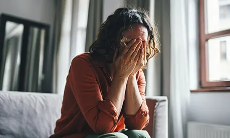 A model poses as a woman covering her face while sitting in a living room, conveying feelings of sadness or emotional stress.
