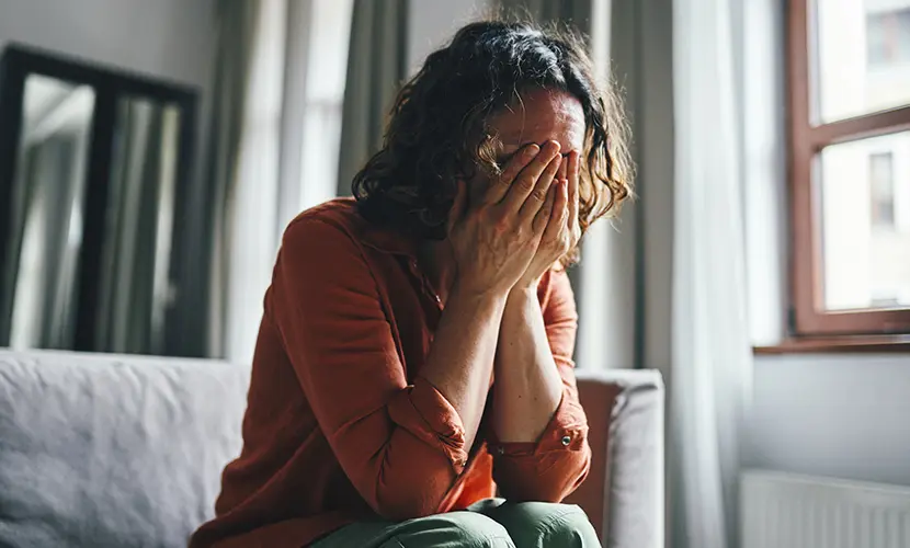 A model poses as a woman covering her face while sitting in a living room, conveying feelings of sadness or emotional stress.