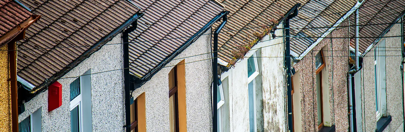 Row of attached houses with sloped, tiled roofs and weathered exterior walls, featuring chimneys, windows, and overhead utility wires.