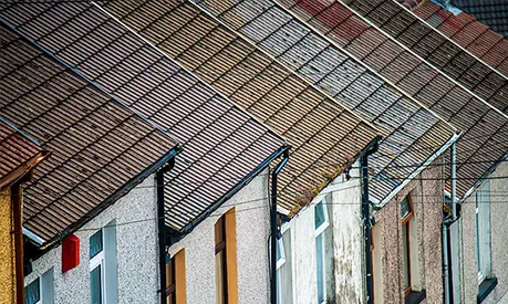 Row of attached houses with sloped, tiled roofs and weathered exterior walls, featuring chimneys, windows, and overhead utility wires.