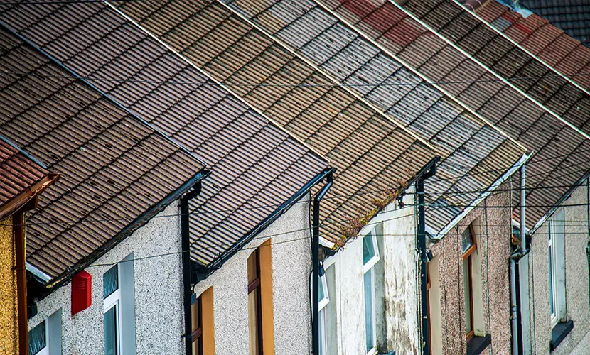 Row of attached houses with sloped, tiled roofs and weathered exterior walls, featuring chimneys, windows, and overhead utility wires.