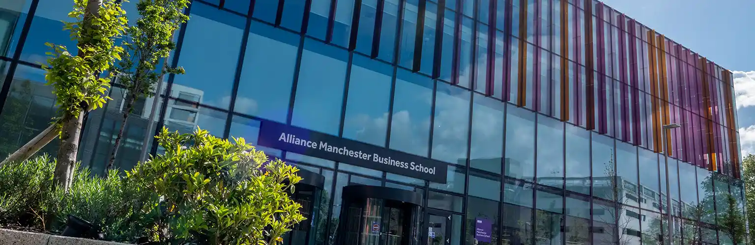 Exterior view of Alliance Manchester Business School building with glass fa&ccedil;ade and colorful vertical panels, surrounded by greenery on a sunny day.