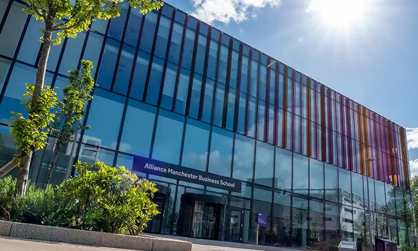 Exterior view of Alliance Manchester Business School building with glass façade and colorful vertical panels, surrounded by greenery on a sunny day.