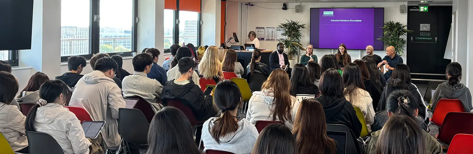 An event with rows of chairs full of students and a panel of five people speaking at the front of the room.