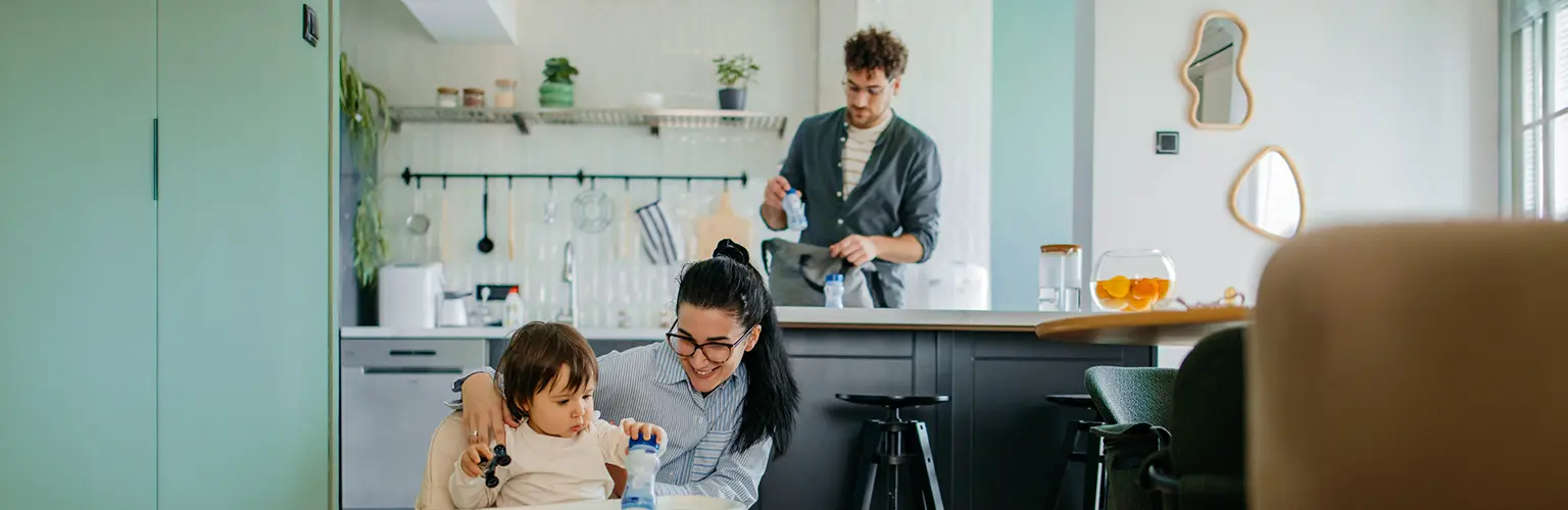 Mother caring for a young child in a high chair while the father prepares items in the kitchen behind them.