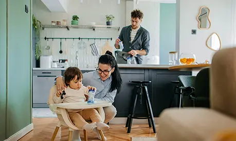 Mother caring for a young child in a high chair while the father prepares items in the kitchen behind them.