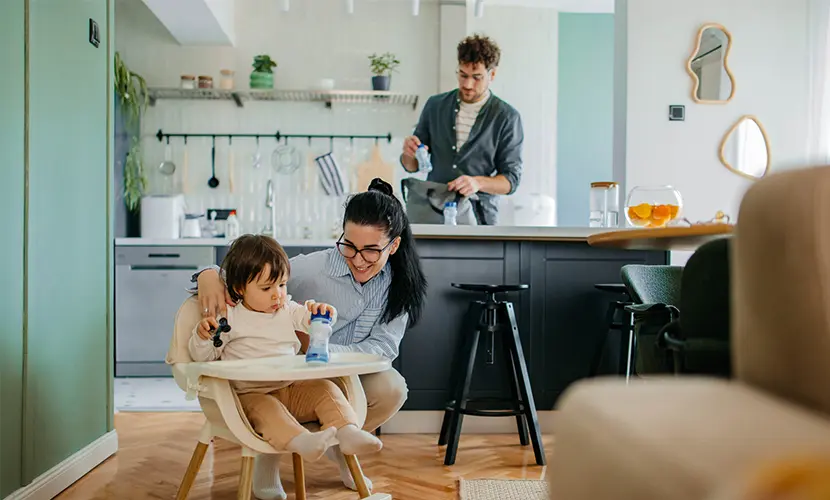 Mother caring for a young child in a high chair while the father prepares items in the kitchen behind them.