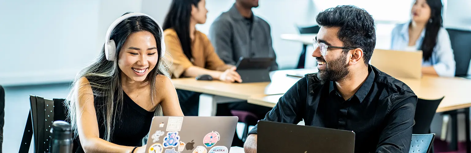Two students working together on laptops in a seminar room.