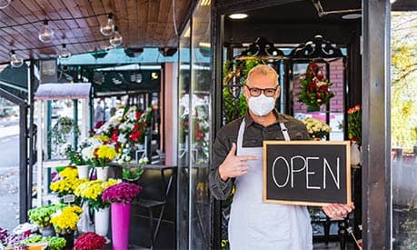 florist in a mask holding up a chalk board that says 'open'