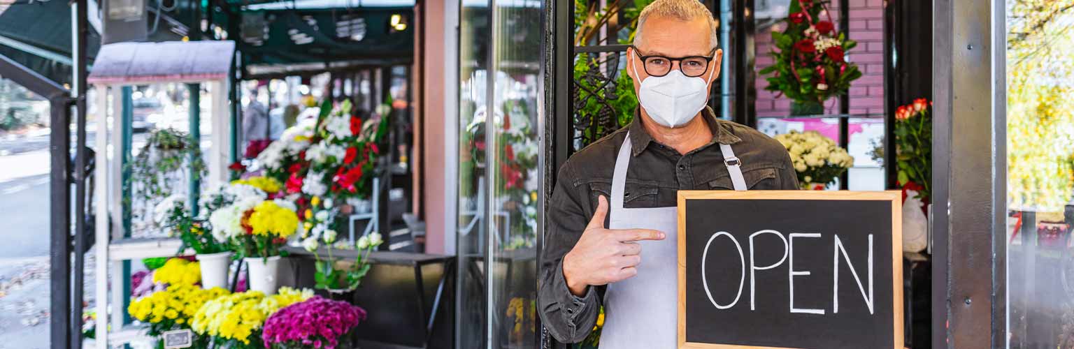 florist in a mask holding up a chalk board that says 'open'