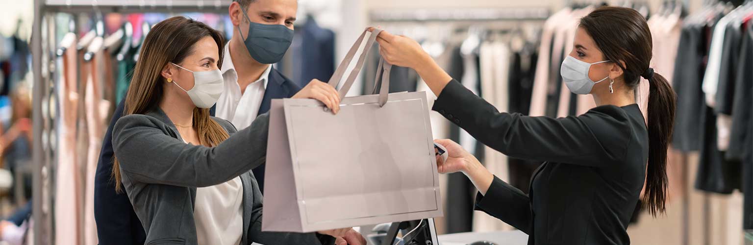 A female retail worker handing over a bag of shopping to a female customer with a facemask on.