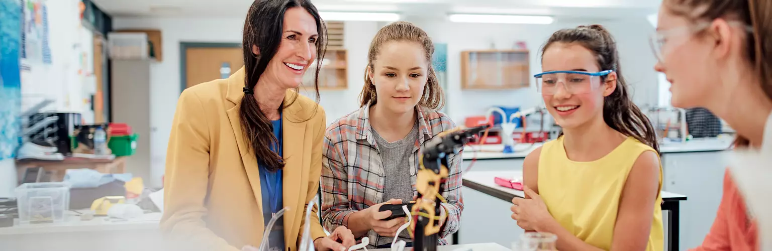 A female teaching helping younger female students with a robot arm on a desk
