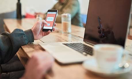 A person holding a smartphone on a cafe table with a laptop in the background