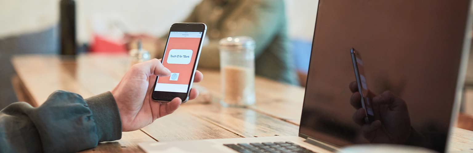 A person holding a smartphone on a cafe table with a laptop in the background