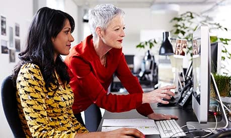 An older employee mentoring a younger employee at her desk