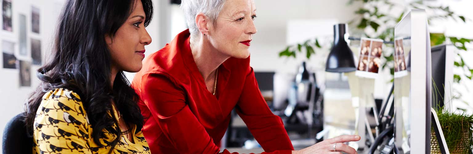 An older employee mentoring a younger employee at her desk