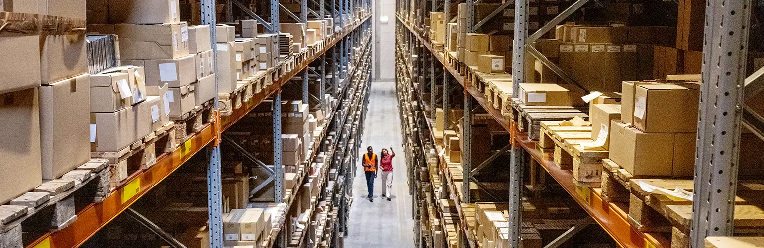 Two people walking through an aisle in a warehouse filled with cardboard boxes and other materials