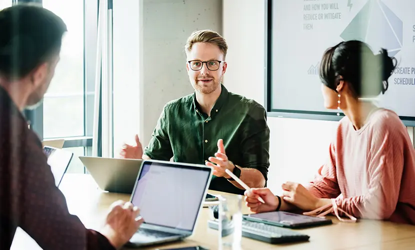 A group of three professionals sat around a table with laptops discussing a group project