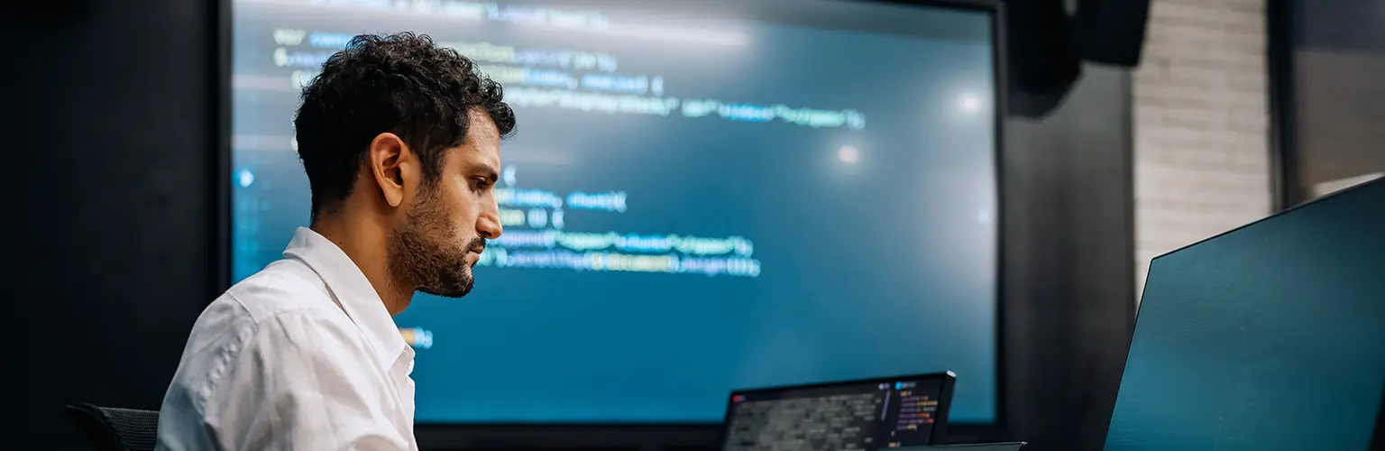 A professional male sitting at a computer which has lots of numbers on the screen
