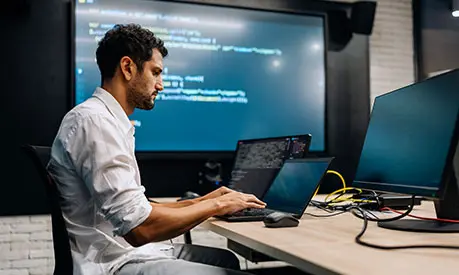 A professional male sitting at a computer which has lots of numbers on the screen