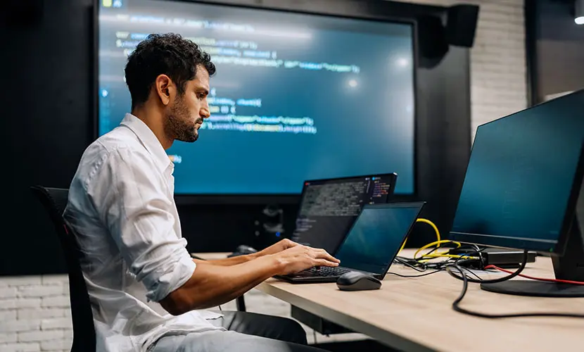 A professional male sitting at a computer which has lots of numbers on the screen