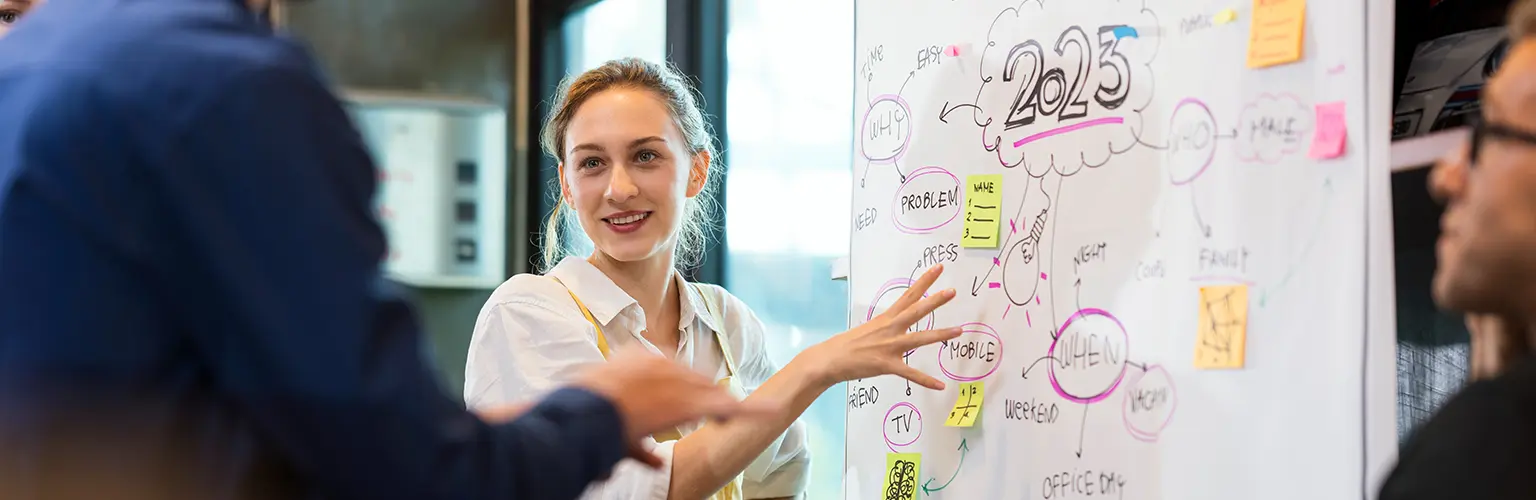 A professionally dressed female standing next to a white board pointing to written ideas in front of her peers
