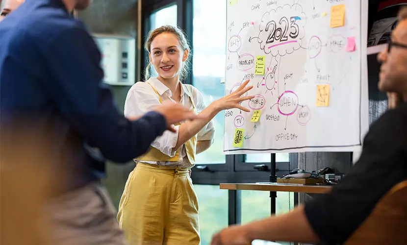 A professionally dressed female standing next to a white board pointing to written ideas in front of her peers