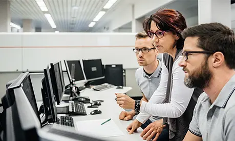 Mature female programmer guiding two engineers in a clean control room of a large factory.