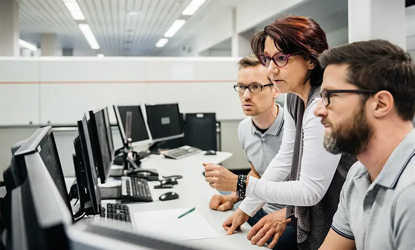 Mature female programmer guiding two engineers in a clean control room of a large factory.