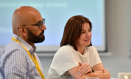 A male and a female dressed in professional clothing talking to other class mates across the table who are out of the frame
