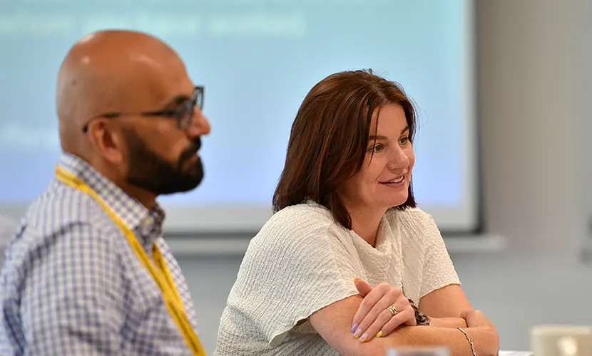 A male and a female dressed in professional clothing talking to other class mates across the table who are out of the frame