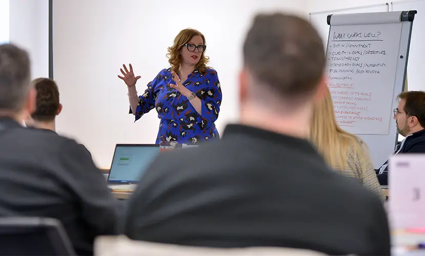 A female academic standing at the front of a classroom delivery a session to professional delegates
