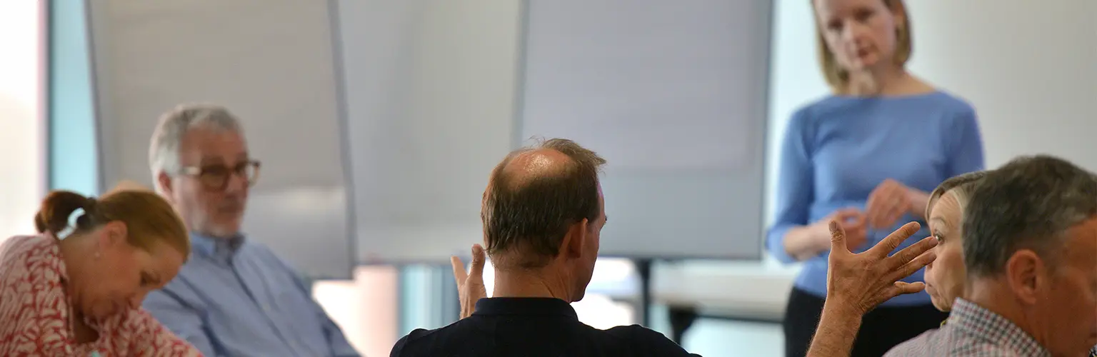 A group of professionals sitting in a classroom environment, with out man speaking to the academic at the front of the classroom