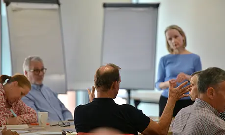 A group of professionals sitting in a classroom environment, with out man speaking to the academic at the front of the classroom