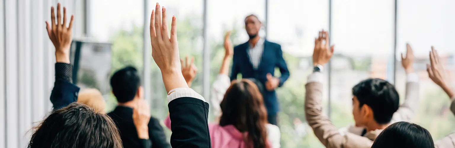 A group of delegates in a classroom with majority of people with their hand up in the air to answer a question.