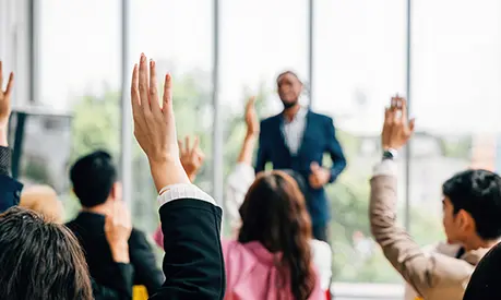 A group of delegates in a classroom with majority of people with their hand up in the air to answer a question.
