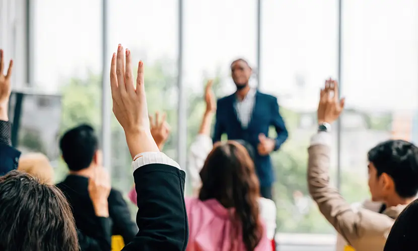A group of delegates in a classroom with majority of people with their hand up in the air to answer a question.