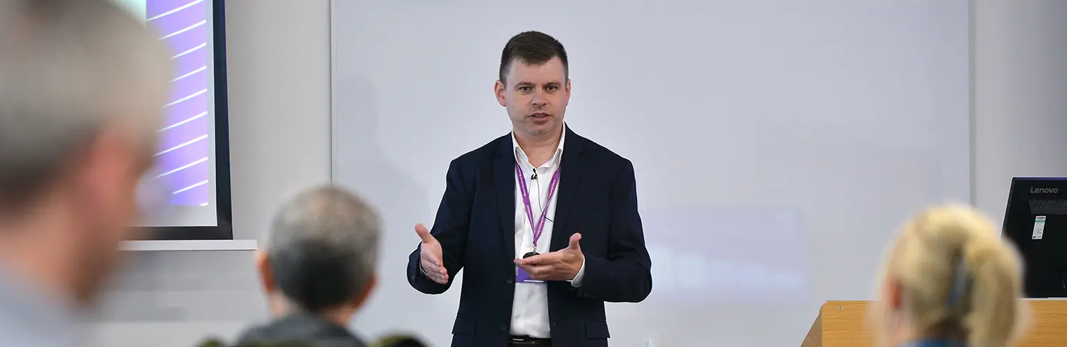 A professional academic stood at the front of a classroom delivering a session to a room of senior leaders