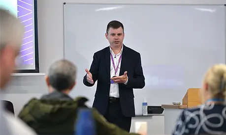A professional academic stood at the front of a classroom delivering a session to a room of senior leaders