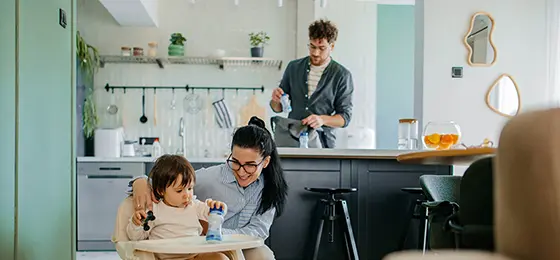 Mother caring for a young child in a high chair while the father prepares items in the kitchen behind them.